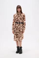 Vestido tipo camisa de corte holgado, con cuello clásico, cierre frontal de botones y mangas largas. Presenta un estampado de hojas en tonos marrones y beige.