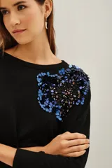 Sweater negro de punto con cuello redondo y mangas largas. Presenta una aplicación decorativa de lentejuelas y pedrería en tonos azules y oscuros sobre el hombro izquierdo.