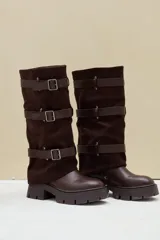 Bota alta de caña ancha, combinada en cuero ecológico y gamuza color marrón, con suela track gruesa y tres tiras horizontales con hebillas metálicas.