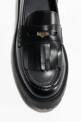 Mocasines de cuero sintético negro con suela chunky, detalles de flecos en el empeine, tira con aplique metálico dorado y costuras pespunteadas.