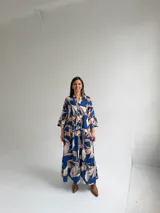 Vestido largo azul con estampado de hojas en tonos beige y blanco, escote en V con botones frontales, mangas largas amplias y diseño escalonado.
