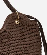 Bolso de playa tipo canasta, tejido en fibra de papel color marrón oscuro, con forma redondeada y abertura superior curva. Cuenta con asas trenzadas de material sintético marrón oscuro y detalles de flecos colgantes a los lados.