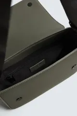 Bandolera verde militar con acabado engomado y textura geométrica. Tiene un compartimento principal con cierre de solapa y botón a presión, un bolsillo interior con cremallera y un asa de hombro ajustable con monedero.