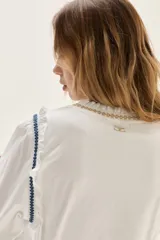 Blusa blanca de algodón con cuerpo de jersey y mangas de popelina. Presenta detalles de punto smock en puños y cuello, adornados con delicados bordados en azul y dorado.