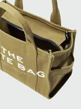 Bolso tipo tote de Marc Jacobs, confeccionado en lona resistente color beige. Presenta el logo estampado en contraste en el frente, doble asa superior y cierre superior con cremallera.