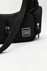 Cartera tipo shoulder color beige, de diseño utilitario y formato curvo. Confeccionada en material liviano y resistente, cuenta con correa regulable para llevar al hombro o cruzada. Presenta múltiples bolsillos con cierre en el frente y laterales, y un parche con el logo "Piece & Cake" en el frente.