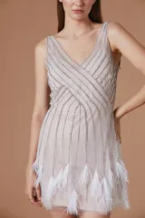 Vestido corto de fiesta color nude, con escote en V y tirantes anchos. Presenta una capa superior de tul transparente decorada con líneas verticales de strass plateado y terminación en el ruedo con plumas blancas.