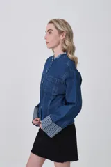 Campera corta de denim azul oscuro, con cuello mao deshilachado y botones plateados frontales. Las mangas largas tienen puños anchos con estampado de rayas verticales en tonos celestes y blancos.