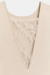 Vestido midi de color beige, con cuello redondo y diseño sin mangas. Presenta un cuerpo liso de punto con bajo asimétrico y una falda inferior confeccionada en encaje tono sobre tono.