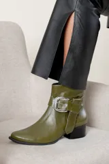 Bota corta de cuero color verde oliva, con taco cuadrado bajo y punta redondeada. Presenta una tira decorativa con hebilla metálica plateada en el tobillo.