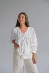 Blusa blanca de corte holgado con cuello en V, cierre frontal con botones y detalles de alforzas y encaje en el pecho. Presenta mangas largas con puños rectos.