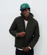 Campera puffer de hombre color verde militar, con capucha integrada, cierre frontal por cremallera y bolsillos cargo frontales. Presenta un diseño acolchado con acabado mate y ajuste en las mangas.