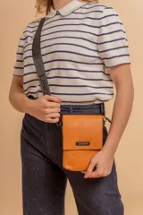 Phonebag pequeña color mostaza, confeccionada en cuero sintético, con solapa frontal y correa ajustable con logo de la marca.