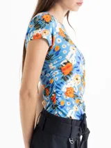 Top de punto color azul con estampado floral en tonos naranja, blanco y verde. Tiene escote redondo, mangas cortas y frunces laterales que estilizan la figura.