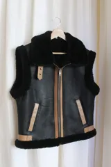 Chaleco estilo bomber de cuero negro con interior y detalles de piel sintética en color crema. Presenta cierre frontal con solapa de cuero en tono tostado y mangas de tejido de punto color crema.
