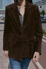 Blazer de piel 100% color marrón oscuro, con cuello solapa, manga larga con hombreras y bolsillos delanteros con solapa. Presenta pespuntes decorativos en contraste y cierre frontal con un botón.