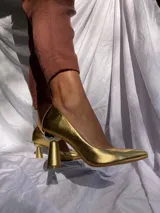 Zapato de tacón alto color dorado, con punta fina y taco con forma geométrica. Capellada, forro y plantilla 100% de cuero.