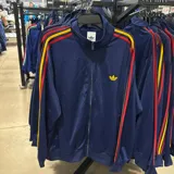 Campera deportiva Adidas Originals de color azul marino con cierre frontal completo. Presenta cuello alto, el icónico logo del trébol en amarillo en el pecho y las clásicas tres tiras en los hombros y mangas en colores rojo y amarillo.