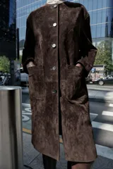 Abrigo marrón de gamuza con cuello redondo, manga larga, bolsillos delanteros y cierre frontal con botones.