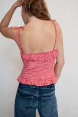 Top de escote redondo y tirantes finos con lazada, con estampado de cuadros vichy en rojo y blanco. Presenta frunces en el pecho y detalle de volados en el ruedo.