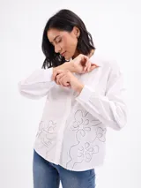 Camisa blanca de manga larga con cuello camisero, cierre frontal de botones y detalles bordados con pespuntes en la parte inferior.