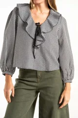 Blusa de manga larga con estampado de cuadros vichy en blanco y negro. Presenta un cuello con volados amplios y un detalle de lazo negro en el centro del escote.