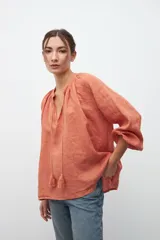 Camisa de corte holgado en color ocre, con escote en V y cordones con borlas decorativas. Presenta mangas largas abullonadas con puños elásticos y un diseño de textura rústica.