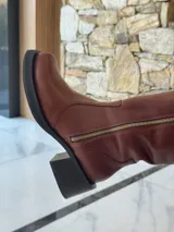 Bota de caña alta estilo biker, confeccionada en cuero color chocolate. Presenta un cierre metálico lateral que recorre toda la caña, estribo y cadena decorativa desmontables, y taco cuadrado con plataforma.