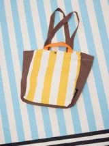 Bolso tipo tote con diseño de rayas verticales en amarillo y blanco, con laterales, base y correas en color marrón. Incluye asas cortas en color naranja y cierre con botón a presión.