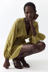Vestido corto oversized de mezcla de viscosa y color verde militar, con escote en V profundo, cordón ajustable en el cuello y mangas globo largas con puños elásticos. Presenta fruncidos en el canesú y sección de punto smock en la cintura con falda de leve vuelo.