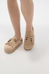 Suecos tipo mocasín color chocolate, confeccionados en material gamuzado. Presentan un diseño con flecos frontales, cordones decorativos con cuentas y una suela de plataforma plana de goma.