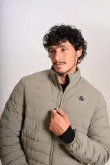Campera puffer de corte clásico en color beige, con cierre frontal, cuello alto y puños elásticos. Presenta un diseño acolchado horizontal y un pequeño logo bordado en el pecho.