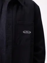 Camisa de gabardina color verde oliva, con cuello clásico, cierre frontal de botones y dos bolsillos frontales con parche. Incluye un pequeño parche con el logo de la marca en el bolsillo izquierdo.