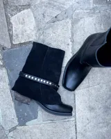 Botas de cuero vacuno color negro, con diseño versátil que incluye una polaina desmontable y un estribo decorativo con tachas metálicas. Presentan un taco bajo y punta redondeada.