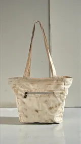 Cartera tipo tote de cuero color natural con bolsillo frontal con solapa metalizada en tono dorado, detalles pintados a mano en la base, flecos decorativos y doble asa de hombro.
