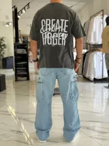 Remera oversize de color gris con efecto gastado (acid wash). Presenta un diseño de tipografía en la parte frontal y un gran estampado en la espalda con la inscripción "Create Bigger". Cuenta con mangas cortas y un calce holgado y cómodo.