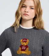 Sweater cropped de punto gris con diseño trenzado, cuello redondo, puños y ruedo con ribetes blancos. Presenta un aplique bordado en el pecho con diseño de osito deportista.