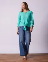 Blusa blanca de tejido texturado con cuello redondo, escote en V con aplique metálico y mangas 3/4 con puños elásticos. El ruedo se ata con un nudo.