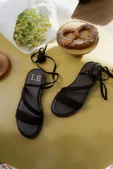 Sandalias planas de cuero color marrón oscuro, con diseño de tiras cruzadas y cordones largos para atar alrededor del tobillo o la pierna. Presentan una suela ligera y flexible.