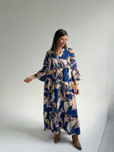 Vestido largo azul con estampado de hojas en tonos beige y blanco, escote en V con botones frontales, mangas largas amplias y diseño escalonado.
