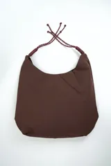 Bolso de hombro color marrón de diseño holgado, con costura central frontal y tiras ajustables de cordón trenzado en tono burdeos con nudos decorativos en los extremos.