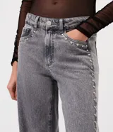 Pantalón de jean color gris lavado, con corte wide leg de piernas amplias y cintura baja. Presenta detalle de tachas metálicas a lo largo de las costuras laterales, cierre frontal con botón y bolsillos funcionales.