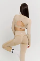 Conjunto deportivo de dos piezas en color beige, compuesto por un top de manga larga con aberturas para pulgares y calzas de tiro alto. Ambas prendas presentan un diseño sin costuras con detalles texturizados y el logo de la marca estampado en el frente del top.