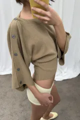 Jersey de punto color beige con cuello barco y mangas anchas tipo capa que presentan detalle de botones a lo largo de los hombros y brazos.