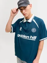 Camiseta tipo polo de manga corta con calce holgado, confeccionada en tejido técnico. Presenta cuello camisero en color blanco, detalles en contraste en hombros y mangas, y estampados gráficos en el frente con el logo de la marca y la inscripción golden hill.