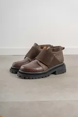 Borcego de caña baja en combinación de cuero liso y gamuza color marrón, con cierre de velcro ajustable en el empeine y suela track dentada negra.
