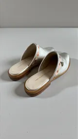 Sandalias chatitas tipo mules, color plateado con detalle de arte en la parte superior y aplique metálico triangular.