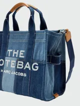 Cartera tipo tote bag Marc Jacobs modelo The Small Traveler Tote, confeccionada en denim con diseño de paneles en distintos tonos de azul. Cuenta con asas superiores, correa de hombro ajustable y extraíble, y el texto "THE TOTE BAG MARC JACOBS" estampado en la parte frontal.
