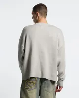 Suéter de punto color gris claro con diseño de escorpión en color blanco tejido en el frente. Presenta cuello redondo, puños y cintura con terminaciones acanaladas, y un corte holgado tipo boxy fit.