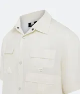 Camisa color crema de corte oversized, con cuello inglés, manga corta y cierre frontal abotonado. Presenta múltiples bolsillos tácticos delanteros.